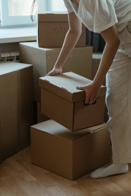 A young woman sitting cross-legged on a wooden floor inside a home surrounded by numerous packed cardboard moving boxes of various sizes, some with red packing tape and labels. She is actively engaged